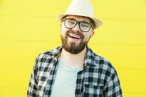 retrato do positivo jovem hipster homem sorridente sobre amarelo parede loja ao ar livre fundo - bonito na moda barbudo cara com chapéu e óculos em pé ao ar livre - moda e na moda juventude pessoas conceito foto