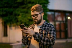 barbudo homem vestindo Óculos é segurando rolagem mensagens de texto dentro dele celular às noite rua. cara chamadas para Táxi dentro a aplicativo dentro tarde cidade foto