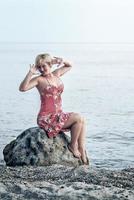 mulher loira feliz em um vestido florido avermelhado. mulher sentada e posar na grande pedra no litoral.