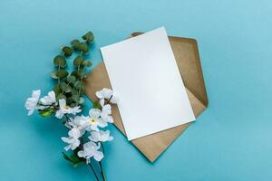 uma kraft papel envelope com uma branco em branco cartão flores em uma azul fundo. cartão postal preparação foto