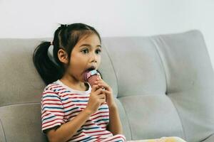 fofa ásia pequeno menina comendo gelo creme em sofá às casa foto
