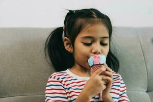 pequeno ásia menina comendo gelo creme enquanto sentado em sofá às lar. foto