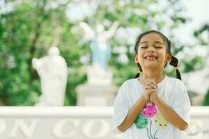 pequeno ásia menina Rezar dentro a parque com uma estátua dentro a fundo foto