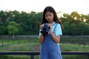 ásia menina usa binóculos para observar a natureza, pássaros, e animais do a nacional parque enquanto acampamento com dela família. com a conceito do Aprendendo lado de fora a Sala de aula natureza estudar, exploração. foto