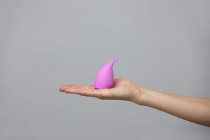 mão de mulher jovem segurando um copo menstrual rosa - fundo cinza. conceito de ginecologia foto