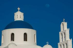 cúpula e Sino torre do a Igreja do panagia platsani e a lua localizado dentro oia cidade às santorini ilha dentro uma lindo cedo Primavera dia foto