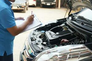 fechar-se homem mecânico inspecionando carro motor. detém papel, verificação e analisando carro motor debaixo a capuz. conceito, ao ar livre carro inspeção serviço. afirmação para acidente seguro. foto