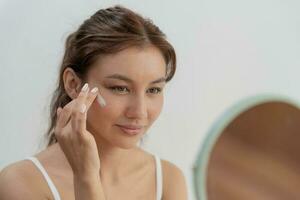 lindo ásia mulher sentar dentro frente do uma espelho. face do uma saudável mulher aplicando creme e inventar. propaganda para pele creme, anti-rugas, bebê face, branqueamento, hidratante, apertar poros sérum foto