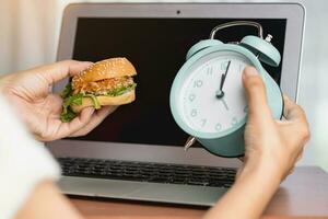 mão segurando uma delicioso Hamburger e alarme relógio enquanto trabalhando foto