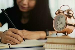 ásia mulher escrevendo em uma caderno com Sino alarme relógio em livros foto