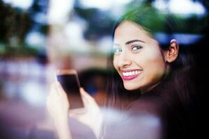 uma lindo jovem indiano mulher sorridente e olhando através uma vidro janela enquanto segurando uma telefone com em branco tela dentro dela mãos foto