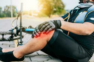 conceito do ciclista com joelho prejuízo ao ar livre. ciclista com joelho dor ao ar livre. masculino ciclista sentado em a calçada com joelho dor foto