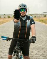 sorridente ciclista mostrando a propaganda em célula telefone, ciclista dentro roupa de esporte mostrando a telefone tela. masculino ciclista em a bicicleta mostrando célula telefone tela ao ar livre foto