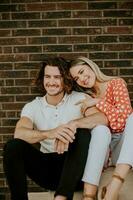 sorridente jovem casal dentro amor sentado dentro frente do casa tijolo parede foto