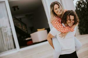 bonito grandes cabelo homem carregando a jovem mulher em dele costas dentro frente do tijolo casa foto