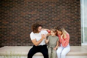 família com uma mãe, pai e filha sentado lado de fora em a passos do uma frente varanda do uma tijolo casa foto