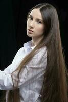 lindo jovem menina com grandes em linha reta cabelo dentro uma branco camisa posando dentro a estúdio em uma cinzento fundo foto