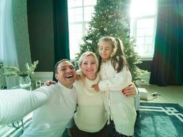 família, feriados, tecnologia e pessoas - sorridente mãe, pai e pequeno menina fazer selfie com Câmera sobre vivo quarto e Natal árvore fundo foto