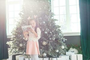 pequeno menina olhando photobook dentro frente do Natal árvore foto
