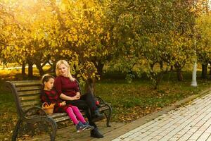 feliz mãe e filha abraçando em uma Banco dentro a outono parque foto