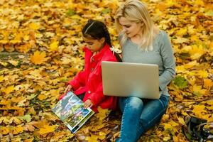 feliz sorridente mãe e criança senta juntos olhando em livro ou tábua pc dentro outono parque foto