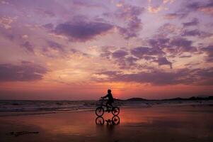 silhueta do pessoa equitação uma bicicleta em uma de praia às pôr do sol foto