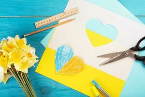 coração recortado em papel e pintado nas cores azul e amarelo e flores amarelas da primavera na madeira de volta. criatividade foto