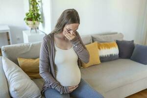 jovem grávida mulher sofrimento a partir de toxicose às lar. maternidade senhora tendo manhã doença cobertura boca com mãos enquanto sentado em sofá dentro vivo sala. mãe Espero por nascer bebê foto