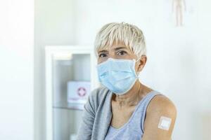 retrato do uma Senior mulher com protetora face mascarar mostrando dela braço com curativo depois de obtendo vacina. maduro mulher sentado depois de recebendo corona vírus vacinação. foto