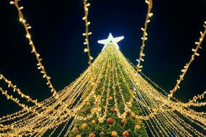 Natal árvore com amarelo guirlandas, decorativo lâmpadas e grande branco Estrela chapéu de coco às noite azul céu foto