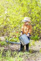 fofa pequeno criança pequena Garoto dentro uma chapéu e borracha chuteiras é rega plantas com uma rega pode dentro a jardim. uma encantador pequeno criança ajudando dele pais crescer legumes. foto