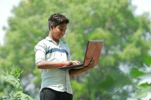 indiano Garoto estudando dentro fazenda, segurando computador portátil dentro mão , pobre indiano crianças foto