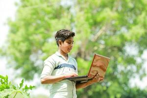 indiano Garoto estudando dentro fazenda, segurando computador portátil dentro mão , pobre indiano crianças foto