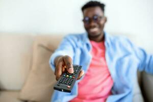 relaxado Preto cara trocando canais em televisão às lar, segurando televisão controlo remoto, sorridente jovem africano homem sentado em sofá dentro vivo sala, assistindo televisão, desfrutando preguiçoso final de semana foto