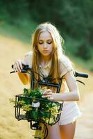 jovem lindo mulher em pé perto amarelo bicicleta com vime cesta cheio do flores dentro floresta foto