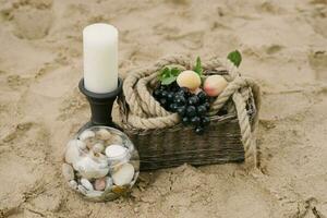 frutas estão dentro uma cesta, uma vela, mar pedras dentro uma vidro vaso. decoração do uma romântico jantar de a mar ou oceano foto