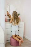 bonita criança menina tintas dela lábios com brilhar ou bálsamo dentro frente do uma espelho dentro dela quarto foto