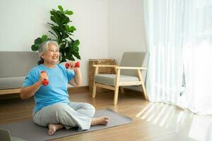 mulher asiática sênior levantando halteres para exercícios e exercícios em casa. mulher madura ativa fazendo exercícios de alongamento na sala de estar. exercite-se ativo e saudável para o conceito mais velho, mais velho e sênior. foto