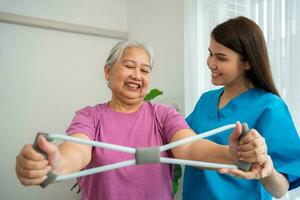 feliz idosos fêmea exercício com borracha expansor com jovem fisioterapeuta para recuperação braço músculos dentro uma fisioterapia Centro. conceito do feliz aposentadoria com Cuidado a partir de uma cuidador e saúde. foto