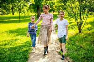 feliz mãe é caminhando com dela filhos dentro parque. elas estão segurando mãos e desfrutando verão dia. foto