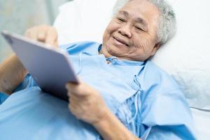 paciente idoso asiático ou idosa segurando um tablet digital nas mãos e lendo e-mails enquanto está sentado na cama na enfermaria do hospital de enfermagem, conceito médico forte e saudável
