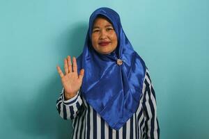 uma amigáveis meia idade ásia mulher dentro uma azul hijab e listrado camisa é sorridente, dizendo oi, e acenando dela mão para cumprimentar uma pessoa, fazer uma Olá gesto enquanto em pé contra uma azul fundo. foto
