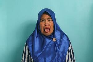 uma descontente meia idade ásia mulher, dentro uma azul hijab e uma listrado camisa, é exibindo uma com nojo expressão Como ela abre dela boca com dela língua visível, enquanto em pé contra uma azul fundo foto