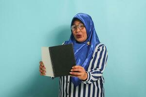 espantado ásia meia idade mulher, vestindo uma azul hijab, Óculos, e uma listrado camisa, detém a aberto livro com uma surpreso expressão enquanto em pé contra uma azul fundo. foto