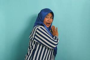 uma assustado meia idade ásia mulher dentro uma azul hijab e listrado camisa suspiros e saltos dentro medo enquanto em pé contra uma azul fundo. foto