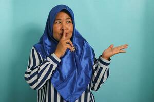 uma meia idade ásia mulher dentro uma azul hijab pontos com dela dedo às a em branco cópia de espaço aparte. ela faz uma silêncio ou estar quieto gesto, dizendo uma segredo venda oferecer, isolado em azul fundo foto