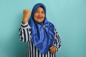 a animado meia idade ásia mulher dentro uma azul hijab e listrado camisa mostra uma Forte gesto de elevação dela braços e flexão dela músculos, sorridente orgulhosamente às a Câmera, isolado em azul fundo. foto