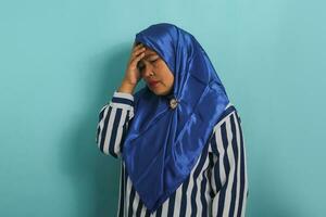 retrato do uma meia idade ásia mulher, vestindo uma azul hijab e listrado camisa, mostra dela sofrimento a partir de uma dor de cabeça, enxaqueca, ou doença, em pé contra uma azul fundo foto