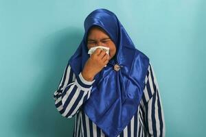 pouco saudável meia idade ásia mulher, vestindo uma azul hijab e uma listrado camisa, é visto sopro dela escorrendo nariz com uma lenço de papel enquanto espirros vencimento para a gripe. cuidados de saúde e médico conceito foto