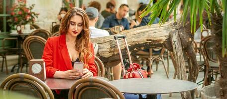 turista jovem caucasiano branco mulher dentro uma vermelho Jaqueta olhando às Smartphone sentado às a mesa dentro cafeteria ao ar livre. foto